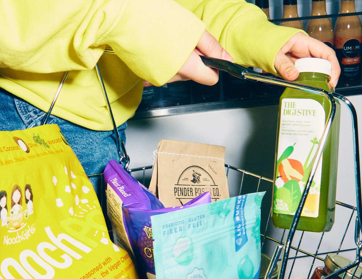 A person in a bright yellow sweatshirt holds a metal grocery basket while holding a bottled green juice labeled 'The Digestive' and other packaged snacks are visible inside the basket.